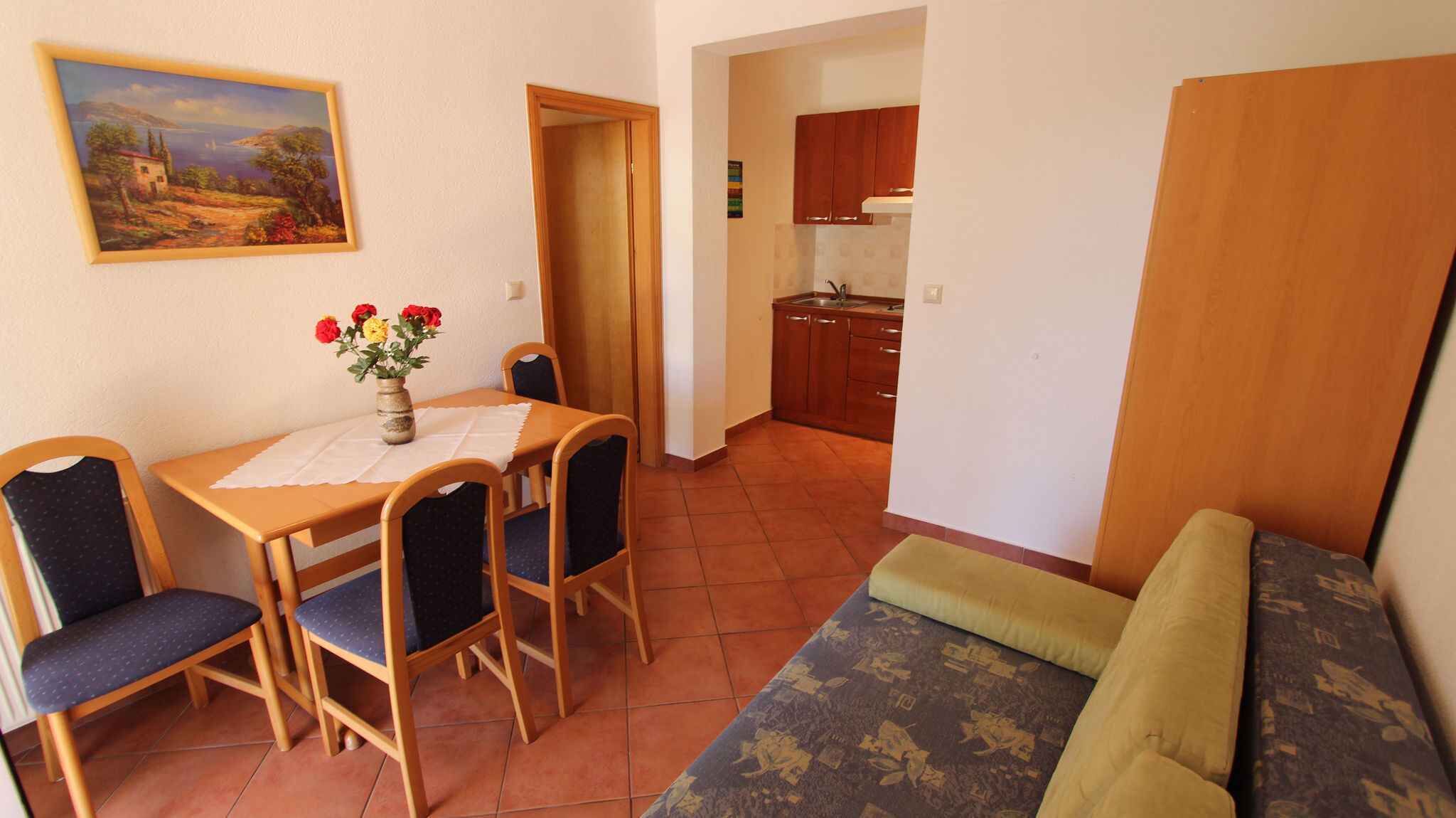 Un comedor con una mesa, sillas y un jarrón de flores. Hay un cuadro en la pared, una cocina al fondo y un sofá con estampado.