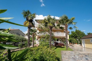 Un bâtiment à deux étages doté d’un balcon, entouré de palmiers et d’un jardin bien entretenu.
