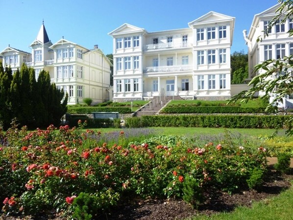A large, multi-story building with a symmetrical facade, a central staircase, and a well-maintained garden with flowering plants.