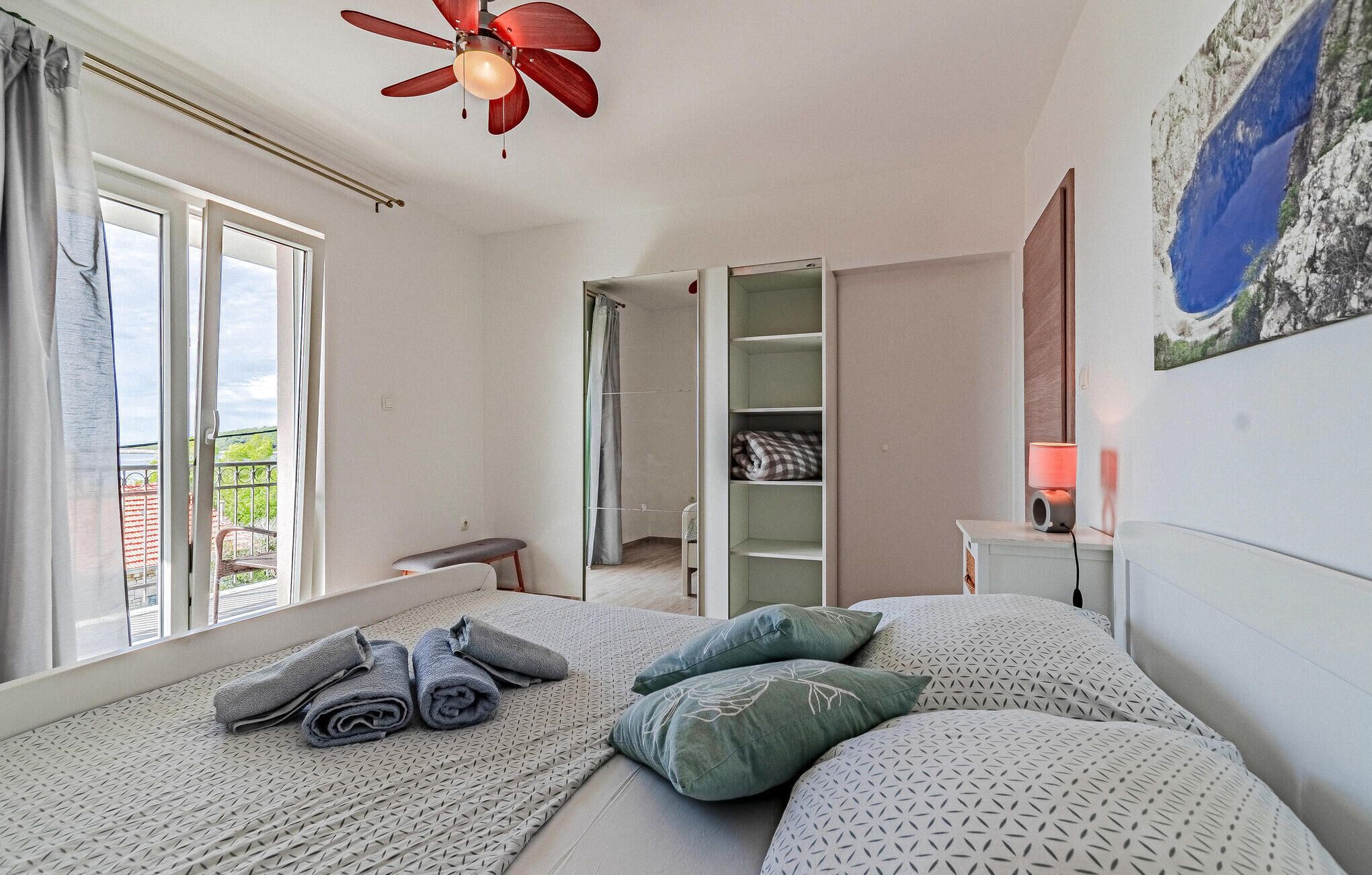 Un dormitorio con cama, ventilador de techo, ventana con cortinas, armario verde y un cuadro en la pared.