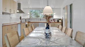 A modern kitchen with white cabinets, a wooden dining table covered with plastic, and a large window providing natural light. - Amazing home in Nexø with sauna (Nexø)