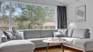 A modern living room with a grey sectional sofa, a round coffee table, and a view of trees through large windows. - Amazing home in Nexø with sauna (Nexø)