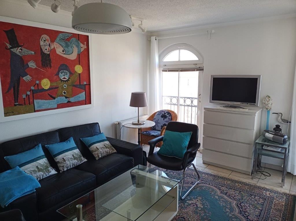 Una sala de estar con un sofá negro, una mesa de centro de vidrio y un cuadro abstracto y colorido. Hay un televisor sobre un mueble bajo, una mesita con una lámpara y un sillón con un cojín color turquesa.