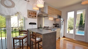 Una cocina moderna con una isla central, sillas de madera, una lámpara colgante y vistas a una piscina a través de la puerta de cristal.