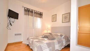 Un dormitorio con cama, cortinas, un televisor montado en la pared y dos cuadros enmarcados.