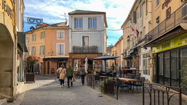 Una calle adoquinada bordeada de edificios, incluyendo un café con terraza y una panadería.