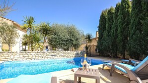 Una piscina con sillas de descanso y una mesa, rodeada de muros de piedra y varios árboles.