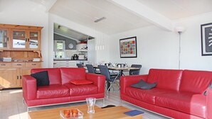 Amplia sala de estar con dos sofás de cuero rojo, una mesa de centro de madera y una mesa de comedor dispuesta para seis personas. Se ve una cocina al fondo.
