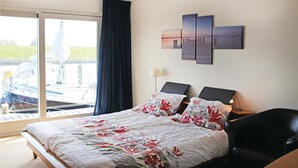 Habitación de hotel con cama, escritorio, silla, lámpara y vista a un barco a través de un ventanal grande.