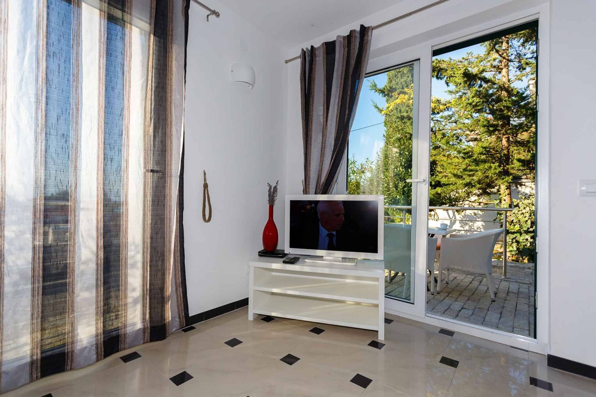 Un soggiorno con una televisione su un mobile bianco, un vaso rosso e una porta scorrevole in vetro che dà su una zona esterna con tavolo e sedie.