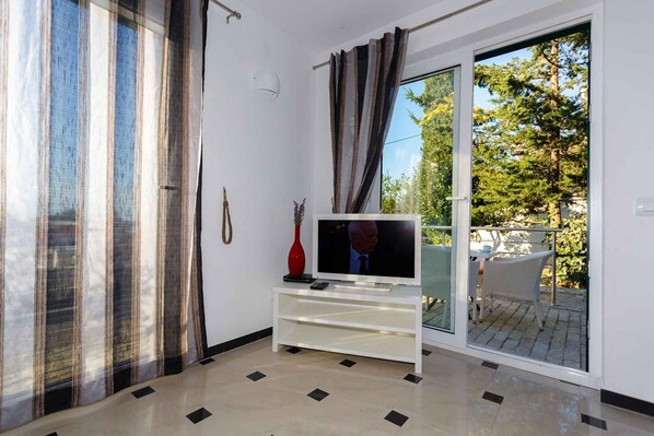 A living room with a television on a white stand, a red vase, and a sliding glass door leading to an outdoor area with a table and chairs.