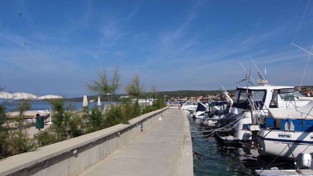 A marina with boats docked, a concrete walkway, and a view of the sea and distant buildings.
