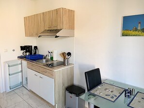 Cocina compacta con armarios de madera, un lavamanos, una cafetera y un refrigerador pequeño. Hay una mesa de vidrio con algunos objetos encima y una silla.