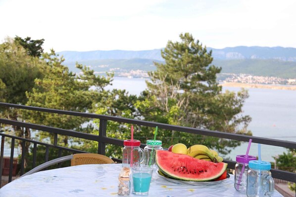 A table with a plate of watermelon and bananas, two drinks, and a bottle of water on a balcony overlooking a lake and mountains.