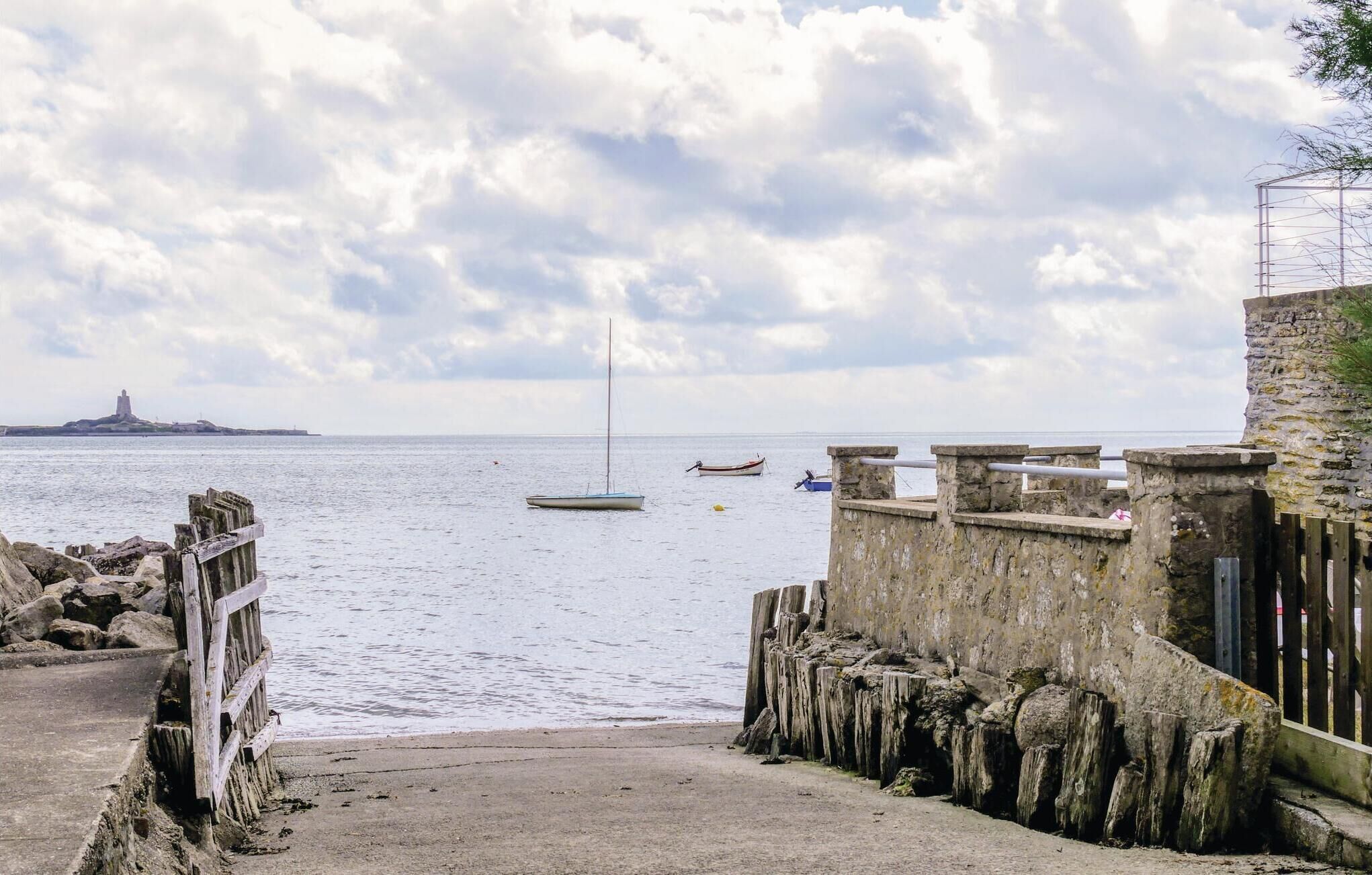 A coastal scene with a stone pier leading to a sandy beach, a small boat anchored in the water, and a distant island with a tower.