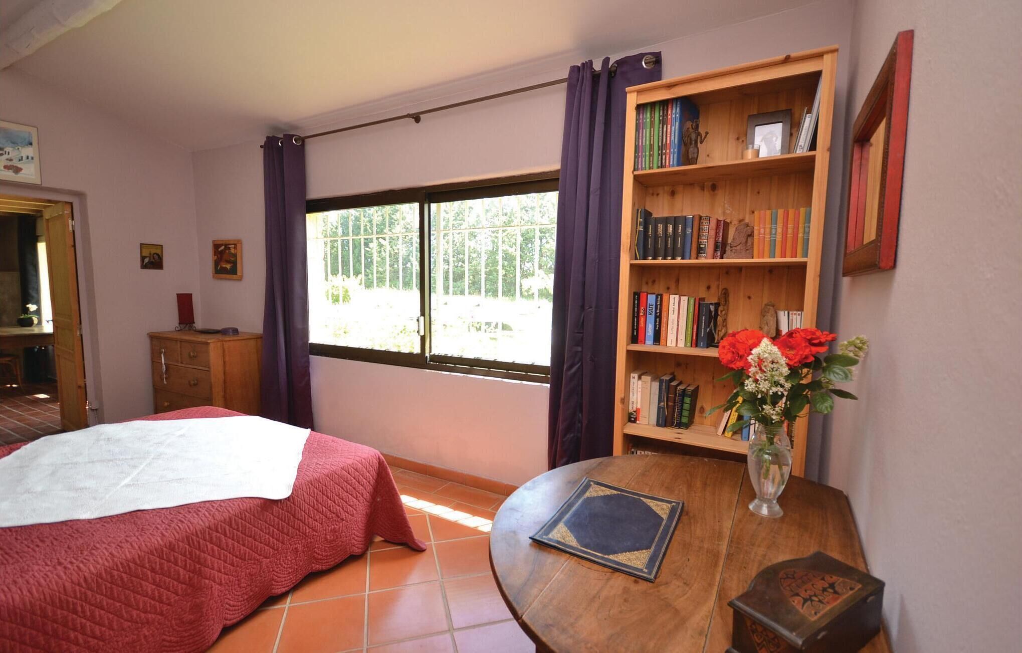 A bedroom with a bed, a wooden bookshelf, a small table with a vase of flowers, and a window with curtains.