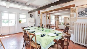 Ein Esszimmer mit einem runden Tisch, gedeckt für vier Personen, umgeben von Holzstühlen. Auf der linken Seite eine weiße Tür mit Fenster, rechts ein Heizkörper und eine Steinwand mit einem gerahmten Bild über dem Tisch.