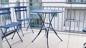 Un balcon avec une table ronde et deux chaises bleues, et en arrière-plan, un bâtiment blanc orné de motifs bleus.