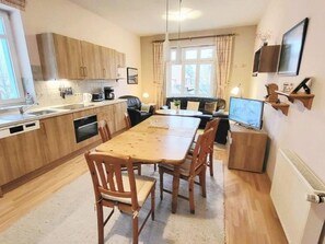 Una cocina compacta con armarios de madera, una mesa de comedor con sillas, un pequeño rincón de descanso con un sofá y un televisor sobre un mueble.