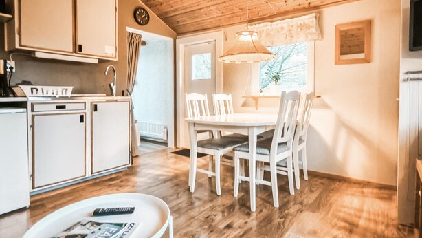 A kitchen with white cabinets, a dining table with chairs, a small round table with a magazine, and a hanging light fixture.