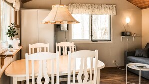 A cozy room with a dining table, chairs, a hanging lamp, a window with curtains, a small shelf with a radio, and a sofa.