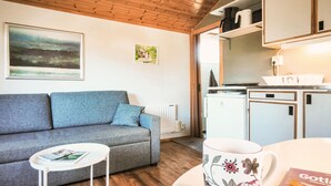 A cozy living room with a grey sofa, a small white table, and a kitchen area with a fridge and cabinets.
