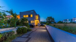 Une maison moderne à deux étages, dotée d’une allée pavée menant à l’entrée, entourée de verdure et d’un petit muret.