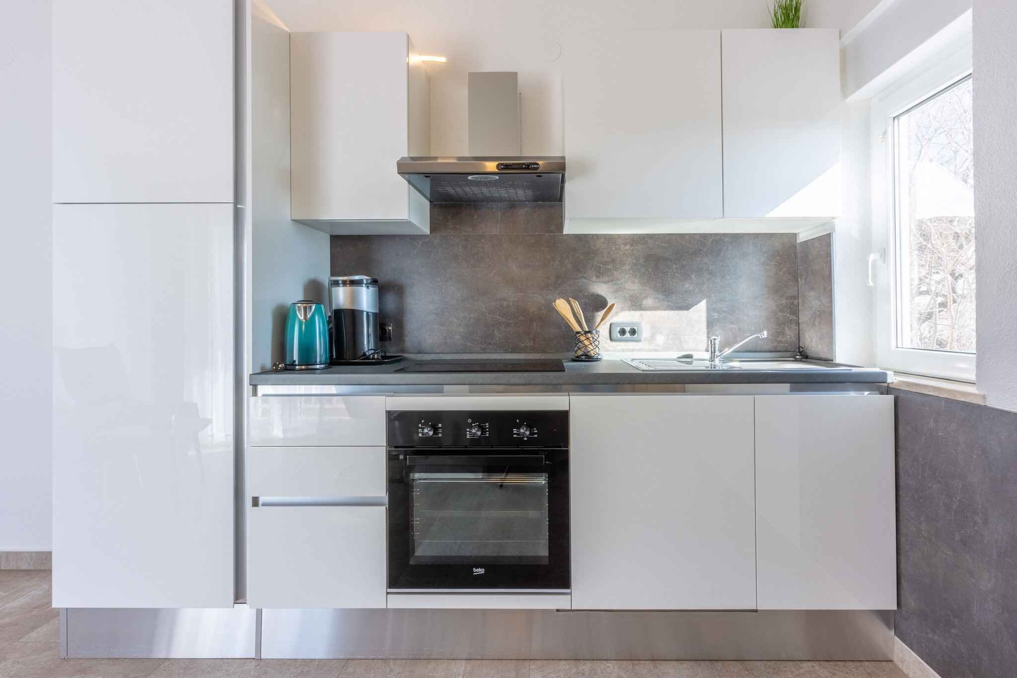Una cocina moderna con armarios blancos, horno empotrado, cafetera y una ventana que proporciona luz natural.