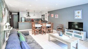 A modern living room with a grey sofa, wooden dining table, and chairs. A TV is on a white stand, and there are wall art pieces. - Stunning home in Lançon de Provence (Lançon de Provence)