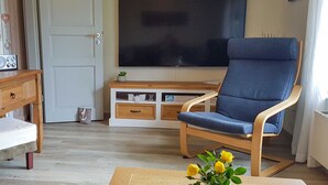 Un soggiorno con TV, una poltrona blu, un tavolino in legno e un vaso con fiori gialli.