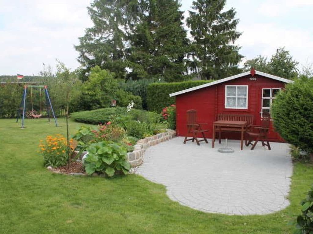 Une maison en bois rouge avec un patio, entourée d’un jardin où se trouve une balançoire.