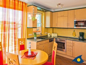 A kitchen with wooden cabinets, a stove, a microwave, and a coffee maker. There is a dining table with chairs and a lamp.