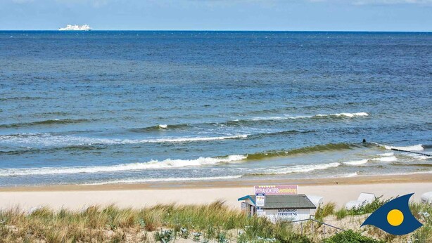 Une plage avec un petit bâtiment portant l’inscription « Location de cabines de plage » et un drapeau avec un cercle jaune.
