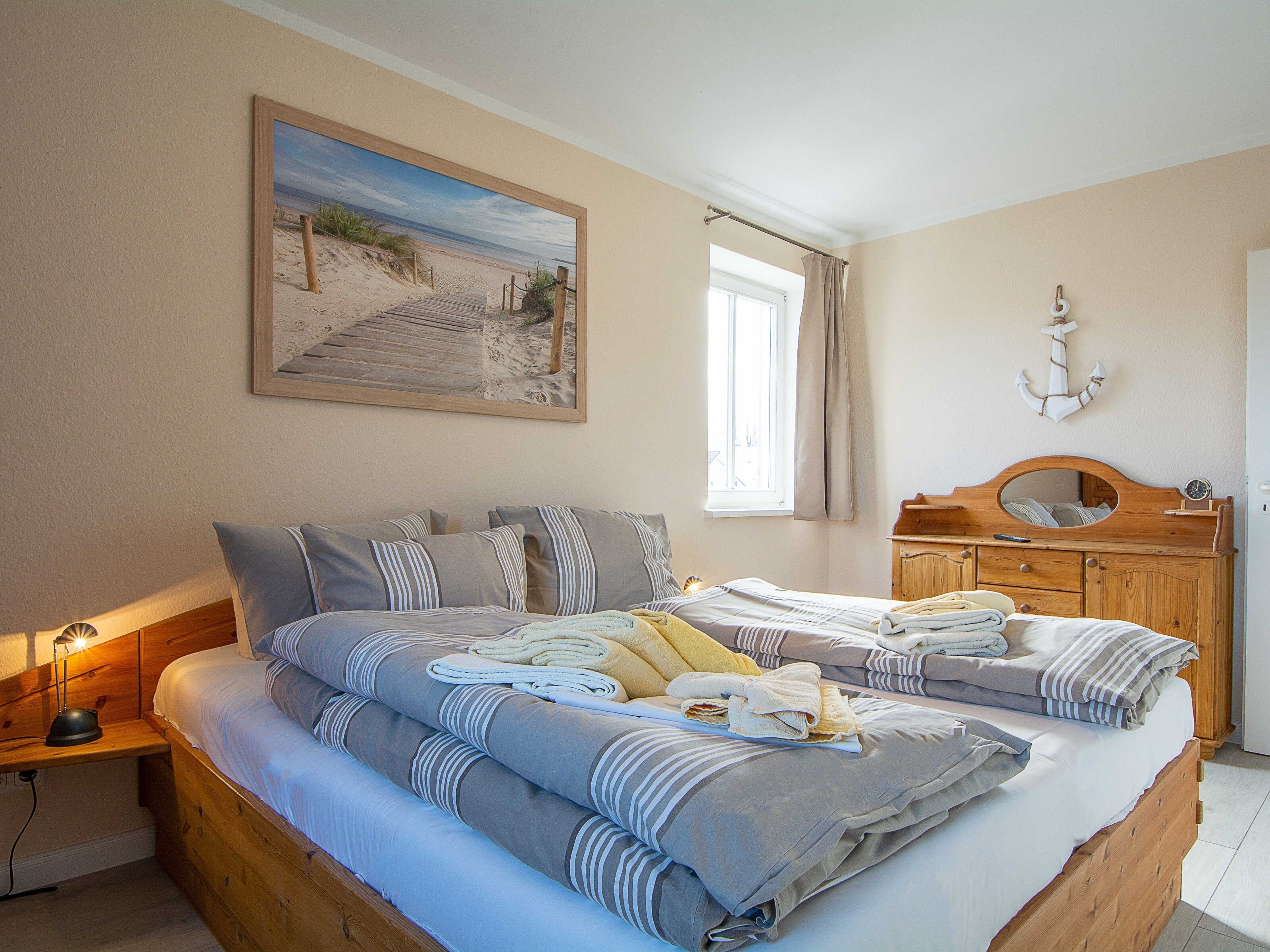 Un lit double avec une literie à rayures, une tête de lit en bois, une table de chevet avec une lampe, une commode en bois avec un miroir et un tableau représentant une scène de plage accroché au mur.