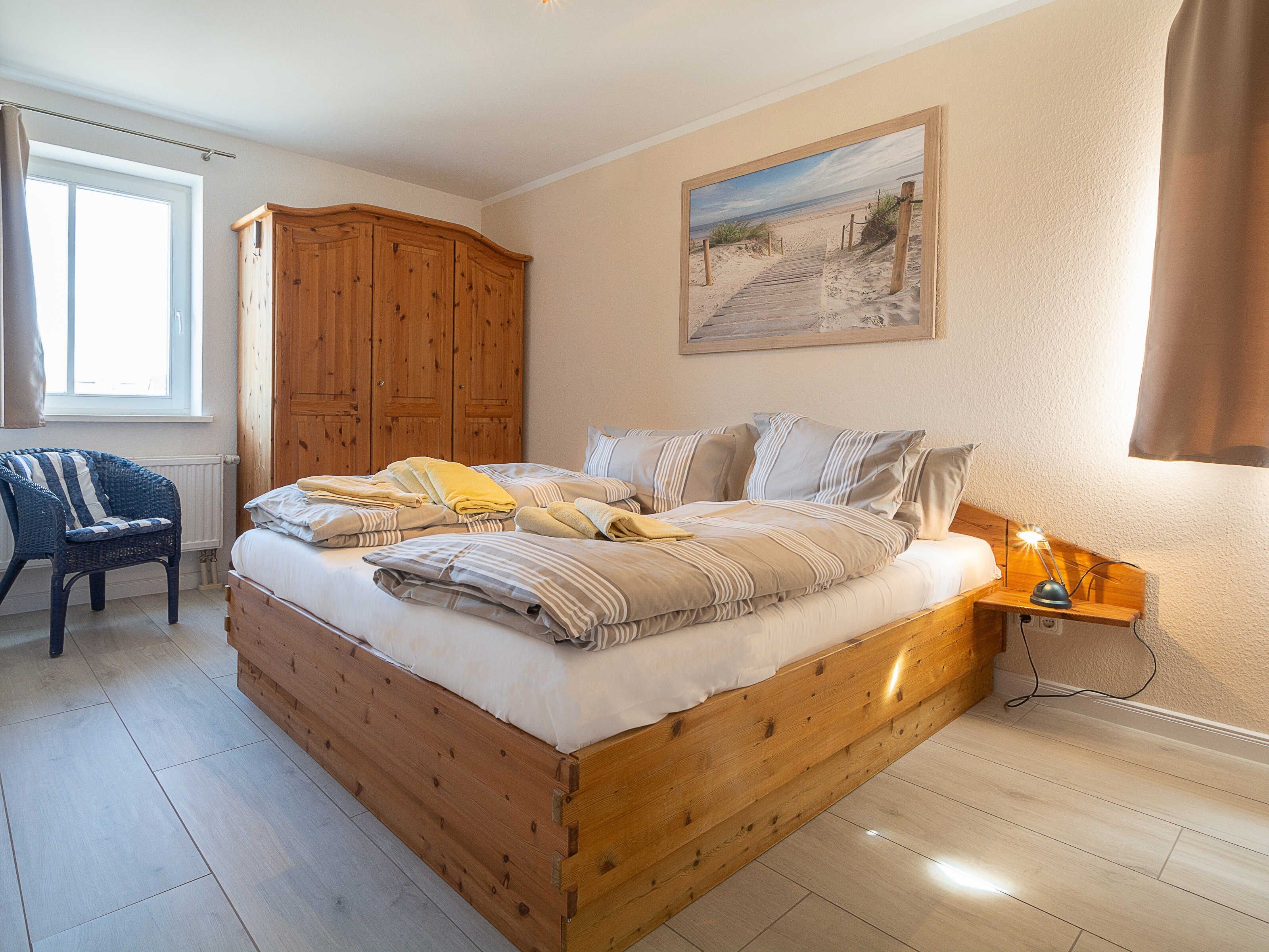 Ein Schlafzimmer mit einem hölzernen Bett, einem Nachttisch mit Lampe, einem Korbstuhl, einem Holzschrank und einem Wandbild mit Strandmotiv.