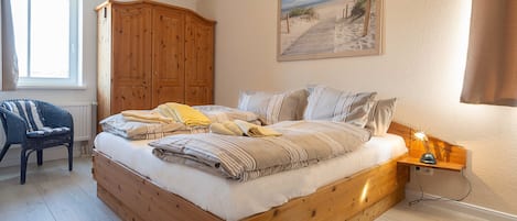 Ein Schlafzimmer mit einem hölzernen Bett, einem Nachttisch mit Lampe, einem Korbstuhl, einem Holzschrank und einem Wandbild mit Strandmotiv.