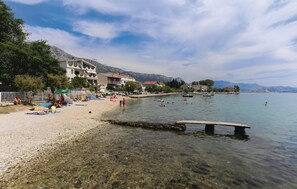 A beach with clear shallow waters, a stone jetty, and buildings along the shore. - Beach front apartment in Kastel Kambelovac (Kastel Kambelovac)