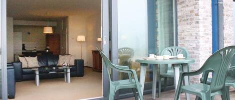 A balcony with a table and chairs, a sofa, and a glass door leading to a living room with a couch and lamps.