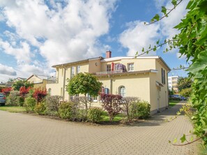 Ein zweistöckiges Gebäude mit Balkon, umgeben von gepflegten Gärten und einer gepflasterten Auffahrt.