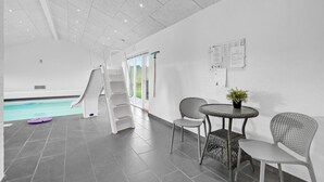 Et moderne poolområde indendørs med en rutsjebane, et bord med stole og en potteplante.