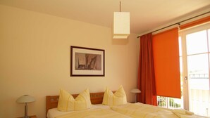 Uma cama de casal com roupa de cama amarela, uma mesinha de cabeceira com abajur, um quadro na parede e uma janela com cortinas laranjas.