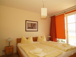 A double bed with yellow bedding, a nightstand with a lamp, a framed picture on the wall, and a window with orange curtains. - LG-08 - Residenz Lindengarten Whg. LG-08th (Kühlungsborn)