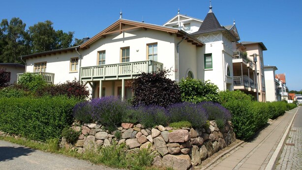 Uma casa grande de dois andares com uma varanda, cercada por vegetação exuberante e um muro de pedra.
