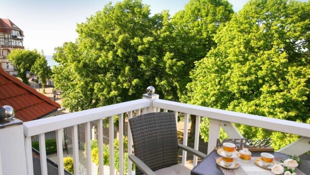 A balcony with a table set for tea, surrounded by lush green trees and a building in the background. - (192) 2-room apartment lake view - Apartment house Baltic Sea beach (Kühlungsborn)