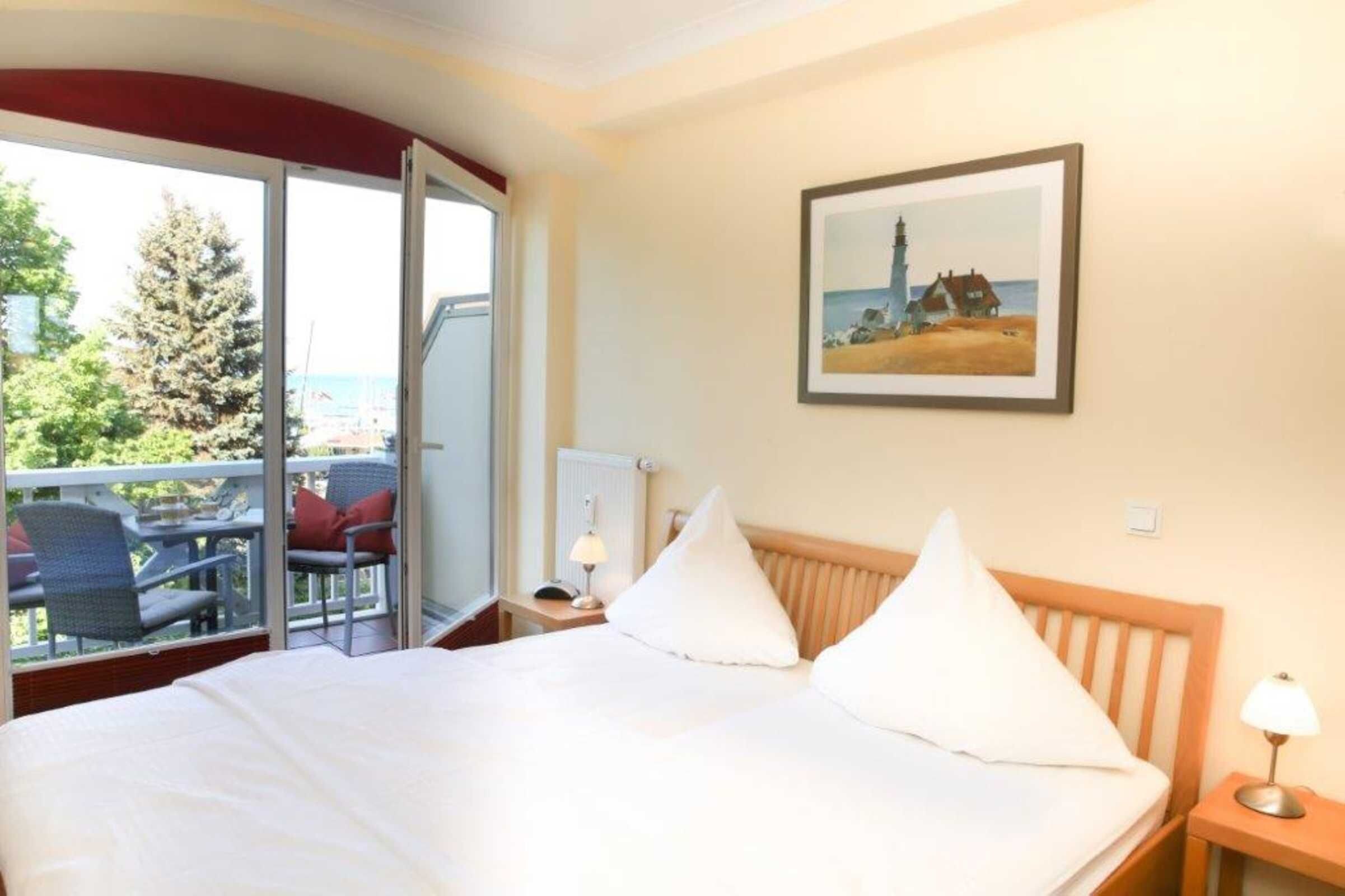 Habitación de hotel con cama, mesita de noche, lámpara, un cuadro en la pared y un balcón con sillas y una mesa.