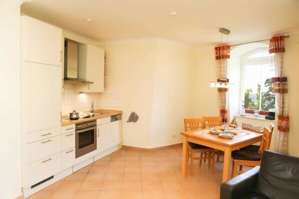 A compact kitchen with white cabinets, a stove, and a dining area with a table and chairs. - (192) 2-room apartment lake view - Apartment house Baltic Sea beach (Kühlungsborn)