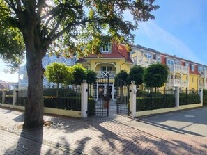 A gated residential area with a yellow building, a tree, and a fence. - (192) 2-room apartment lake view - Apartment house Baltic Sea beach (Kühlungsborn)