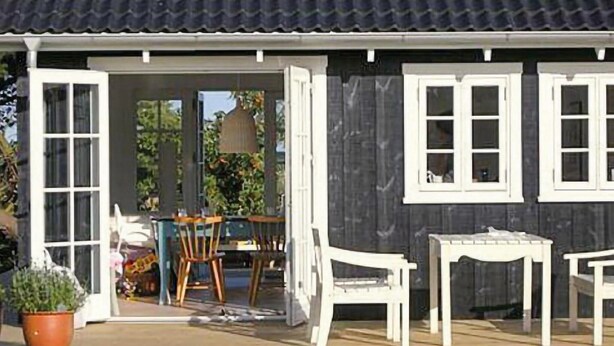 A wooden building with a dark roof, white-framed windows, and a door leading to an outdoor area with a table, chairs, and a potted plant. - Gorgeous home in Sydals with WiFi (Sydals)