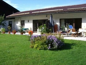 A building with a tiled roof, a covered patio area with outdoor furniture, and a well-maintained lawn with various plants.