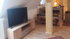 A loft-style room with a flat-screen TV on a wooden cabinet, a dining area with a table and chairs, and a lamp on a desk. - Apartment 3 - Guesthouse Alte Schule (Schwarz)